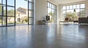 Photographie d'un vaste espace intérieur (salon, cuisine ou showroom) avec un sol entièrement recouvert d'un revêtement de sol de haute qualité (carrelage grand format, béton ciré lissé ou résine époxy mate) de couleur neutre ou grise, parfaitement fini et homogène, photographié depuis un angle qui met en évidence la régularité et la finition professionnelle du sol, avec un éclairage naturel mettant en avant la qualité de surface et la planéité du revêtement.