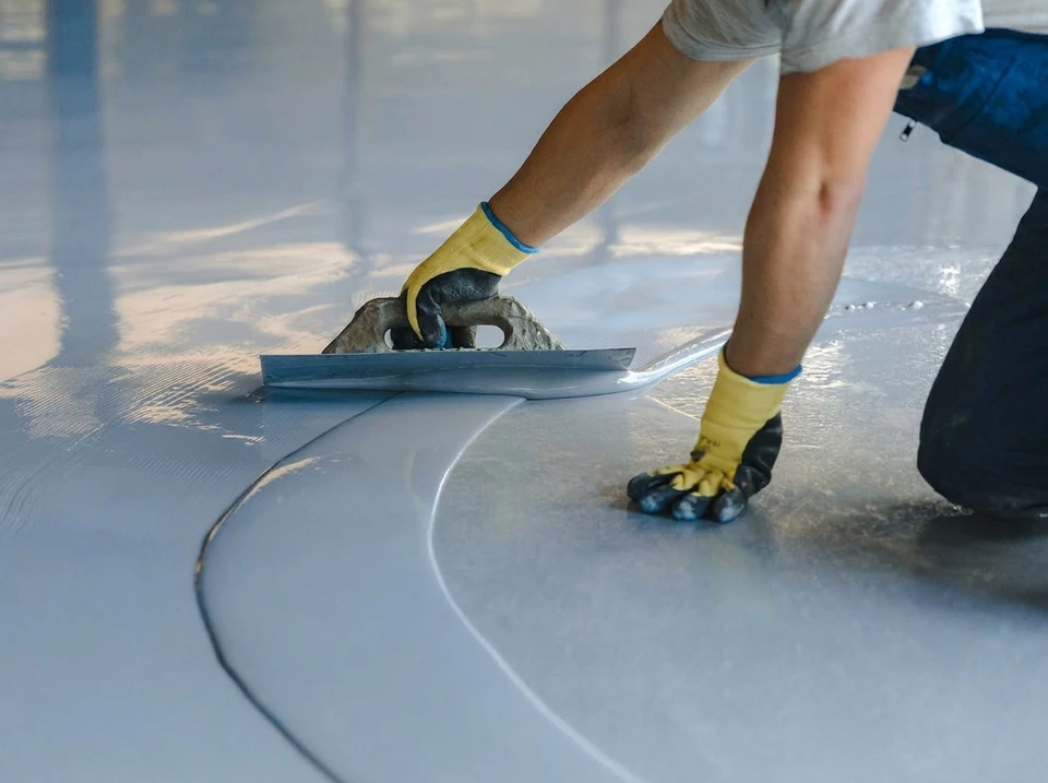 Photographie professionnelle en chantier montrant une équipe de chapistes expérimentés appliquant une chape liquide dans un espace de construction. On voit deux à trois artisans au travail, concentrés sur l'application du matériau au sol, avec des outils professionnels (règle de maçon, perceuse, etc.). L'arrière-plan montre un chantier de construction d'ampleur avec structure bâtie visible. L'image doit transmettre la stabilité, la compétence technique et l'expérience à travers la qualité du travail effectué et la maîtrise professionnelle visible. La photo doit être claire, bien éclairée et de qualité professionnelle, sans texto ou logo visible.
