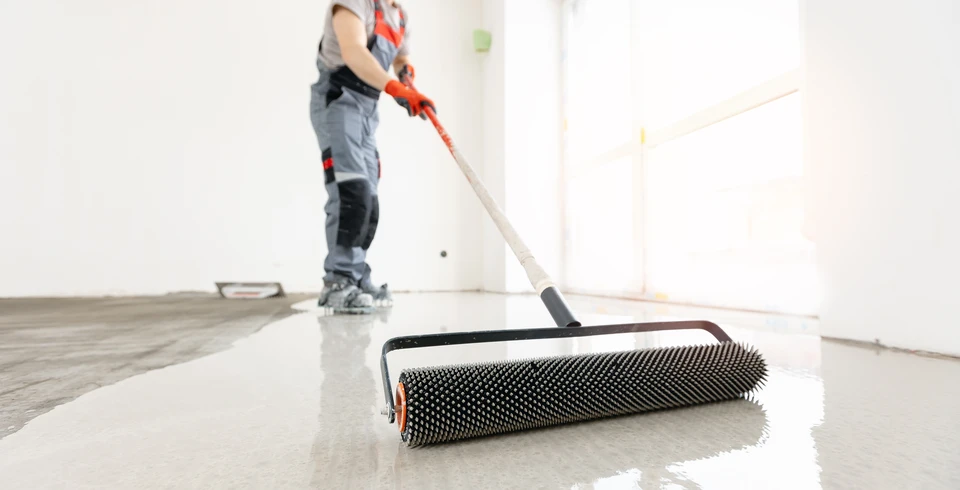 Photographie d'un chantier de travaux de sol en intérieur montrant plusieurs étapes de finition simultanément : au premier plan, un artisan en tenue professionnelle applique une chape liquide grise sur le sol brut, au centre un deuxième ouvrier pose du carrelage ou un revêtement, et en arrière-plan on aperçoit des panneaux d'isolation thermique ou de plancher chauffant. L'ambiance doit être professionnelle, bien éclairée, montrant un chantier structuré et organisé avec du matériel de qualité. Les ouvriers portent des équipements de sécurité appropriés (gilets, chaussures de travail). La composition doit transmettre l'idée d'une prise en charge globale et coordonnée d'un projet complet de sol.