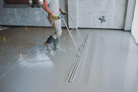 Photographie en gros plan d'un artisan posant du parquet ou un revêtement de sol souple en intérieur. L'artisan doit être en équipement de travail professionnel (gilet de sécurité, bottes). Il applique le revêtement sur une surface préparée en utilisant les outils appropriés. On doit voir clairement les lames de parquet ou le sol souple partiellement posé, avec les matériaux de finition visibles (colle, joints ou underlay selon le type). L'arrière-plan doit montrer un intérieur de chantier avec des zones de travail préparées. La composition doit mettre en évidence le professionnalisme, la technique de pose spécifique au parquet/sol souple et le travail de finition, distinctement différente des scènes de chape liquide ou de carrelage.