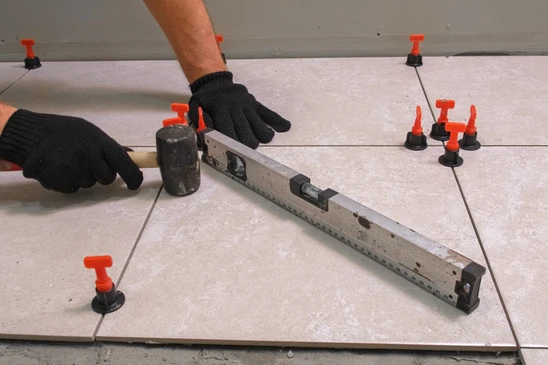 Photographie en gros plan d'un chapiste professionnel en équipement de travail (gilet de sécurité, bottes) effectuant le ragréage et la préparation d'une surface de sol. L'artisan utilise une taloche ou une spatule de ragréage pour lisser et préparer une surface inégale, créant une base uniforme. On voit clairement le composé de ragréage gris en cours d'application sur une surface en béton brut ou inégale, avec le contraste entre les zones préparées (lisses) et les zones non encore traitées. L'arrière-plan montre un chantier intérieur avec une structure de bâtiment visible. La composition met en évidence le professionnalisme, la technique de préparation précise et l'importance de cette étape fondamentale dans la chaîne de revêtement de sol.