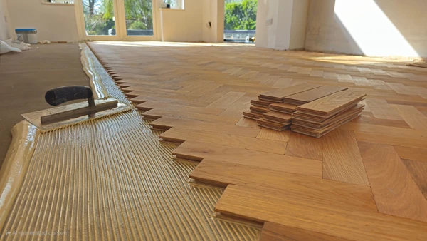 Photographie en gros plan d'un chapiste professionnel en équipement de travail (gilet de sécurité, bottes de protection) installant un système de plancher chauffant sur une surface de chantier intérieur. L'artisan positionne et fixe les éléments chauffants (tubes de pex ou tapis chauffants visibles) sur une surface préparée, potentiellement à proximité d'une pompe de distribution ou d'un système de contrôle du plancher chauffant. On doit clairement voir les tubes ou éléments chauffants en cours d'installation, les fixations, et le contraste entre les zones préparées et les zones non encore équipées. L'arrière-plan montre un chantier intérieur avec une structure de bâtiment visible. La composition met en évidence le professionnalisme, la technique d'installation précise du système de chauffage intégré et l'expertise requise pour cette installation spécialisée.